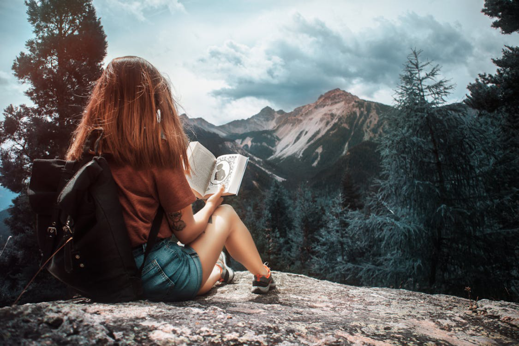 A person reading a fiction book on a hilltop wearing a backpack