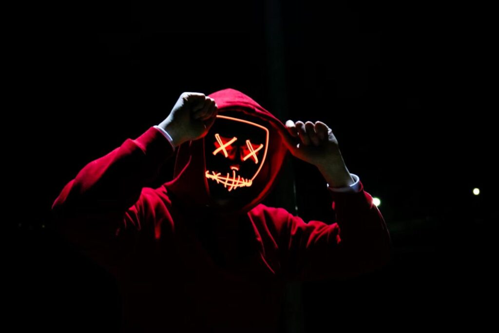 A person wearing a red hoodie and a glowing neon mask with a sinister expression.